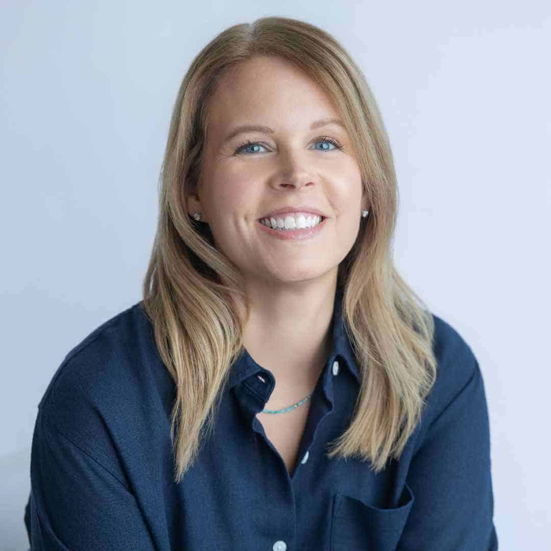 A woman with straight, shoulder-length blonde hair is smiling at the camera. She is wearing a dark blue button-up shirt and a delicate necklace, posed against a light, plain background.