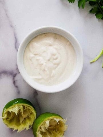 A small white bowl filled with lime crema sits on a marble surface. Beside it are two squeezed lime halves and fresh cilantro sprigs.