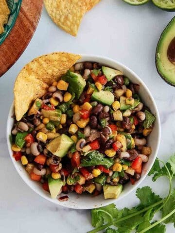 A bowl of texas caviar dip with beans and vegetables, avocado, corn, and peppers, garnished with a tortilla chip. Fresh avocado, cilantro, lime, and more chips are nearby.